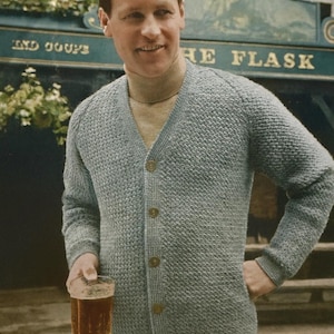 May include: A light blue cardigan with a textured knit pattern, featuring a V-neck and button closure. The man is holding a pint of beer. The pub sign reads "THE FLASK".