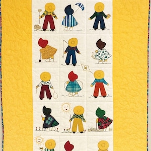 Puede incluir: Un edredón hecho a mano con un patrón repetido de figuras caprichosas. Cada figura tiene una cabeza amarilla en forma de sol y usa diferentes atuendos. Las figuras están dispuestas en una cuadrícula sobre un fondo blanco, sobre un edredón amarillo brillante.