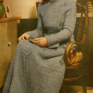 Könnte beinhalten: Ein Vintage-Set aus hellblauem Strick, bestehend aus Pullover und Rock. Das Set hat ein strukturiertes Muster und wird von einer Person getragen, die auf einem Holzstuhl sitzt. Das Outfit hat lange Ärmel und einen hohen Ausschnitt. Der Rock ist lang und gerade.