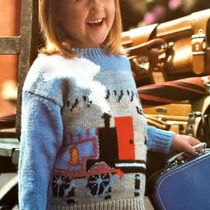 May include: A child's blue and gray knit sweater featuring a train design with red and black accents. The sweater has long blue sleeves and a ribbed neckline and cuffs. The child is holding a blue suitcase.