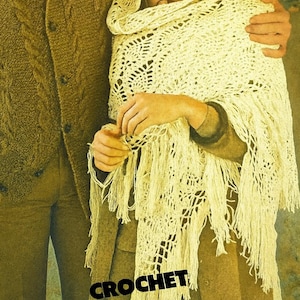Op de afbeelding: Een crèmekleurige gehaakte sjaal met franjes is over de schouders van een persoon gedrapeerd. De sjaal heeft een open ontwerp. Het woord "CROCHET" is in zwarte blokletters gedrukt. De persoon draagt een bruine outfit.