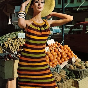 May include: A woman wearing a striped halter dress and a large sun hat. The dress features horizontal stripes in shades of brown, orange, and green. She is standing near a fruit stand with baskets of produce.