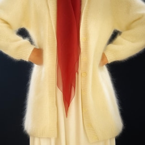 May include: A pale yellow cardigan with a shawl collar and button closure. A red scarf is draped over a white dress. The cardigan has long sleeves and is made of a soft, fuzzy material. The outfit is set against a dark background.
