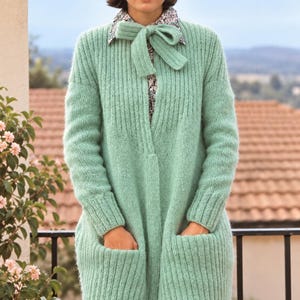 Könnte beinhalten: Eine mintgrüne, langärmelige Strickjacke im Rippenmuster mit weicher, flauschiger Textur, zwei großen aufgesetzten Vordertaschen und einem passenden Strickband am Ausschnitt, getragen über einem gemusterten Hemd mit kleinem, dunklem, filigranem Druck.