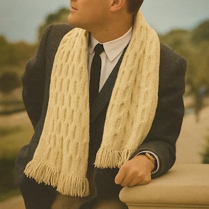 May include: A cream-colored, knitted scarf with a repeating oval pattern and fringe. The scarf is draped over a dark suit and white shirt. The person is wearing a gold watch and leaning on a stone wall.