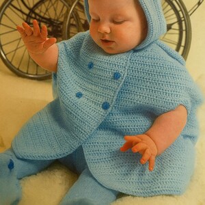 May include: A baby wearing a light blue crocheted outfit, including a hooded cape and matching leggings. The outfit has button closures. The baby is sitting on a white, fluffy surface.