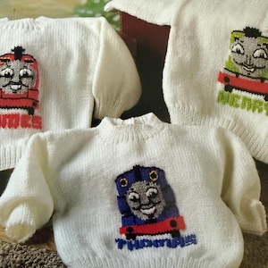 May include: White knit sweaters with colorful embroidered train designs. The sweaters feature different train faces in red, green, and blue, with the name "Thomas" below the blue train. The sweaters are likely for children.