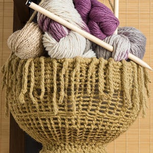 May include: A woven basket filled with yarn in shades of beige, purple, and gray, with wooden knitting needles. The basket has a fringe detail and is suspended from a dark wooden structure, suggesting a craft or hobby theme.