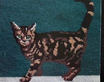 May include: A knitted image of a cat with brown and tan markings on a teal background. The cat is standing with its tail up, and its green eyes are visible. The cat is on a white surface.