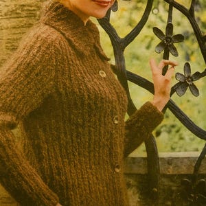 May include: A brown, button-down knit cardigan with a collared neckline. The textured sweater has long sleeves and is made of a thick, warm material. The cardigan is paired with a light-colored bottom. The background includes a decorative metalwork.