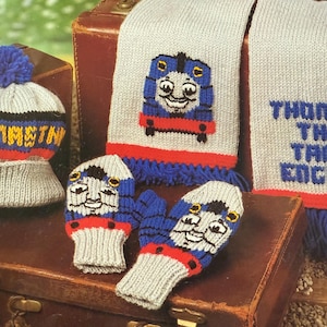 Puede incluir: Un conjunto de punto para niños con un diseño de tren azul. El conjunto incluye un gorro con pompón, una bufanda y manoplas. El gorro y las manoplas tienen una base gris con detalles rojos, azules y amarillos. La bufanda tiene un diseño de tren azul y flecos azules.