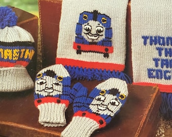 May include: A knitted children's set with a blue train design. The set includes a hat with a bobble, a scarf, and mittens. The hat and mittens have a grey base with red, blue, and yellow accents. The scarf has a blue train design and blue fringe.