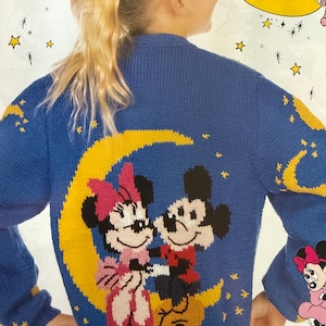 Puede incluir: Un suéter azul tejido con Mickey y Minnie Mouse sobre una luna creciente amarilla. El suéter tiene estrellas amarillas y diseños de luna creciente en las mangas. El texto "Mickey and Minnie instructions begin on page 71" está en la parte inferior.