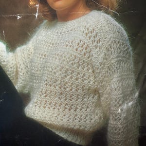 May include: Cream-colored, open-weave sweater with a round neckline and long sleeves. The knit pattern features a delicate, airy design. The sweater appears soft and slightly fuzzy, suggesting a comfortable, warm garment. The overall style is casual and relaxed.