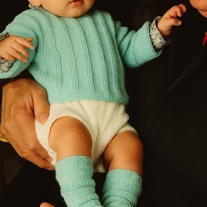 May include: A baby wearing a light blue knitted sweater, matching socks, and white diaper cover. The sweater has a ribbed design. The baby is being held by an adult. The sleeves of the sweater have a floral pattern.