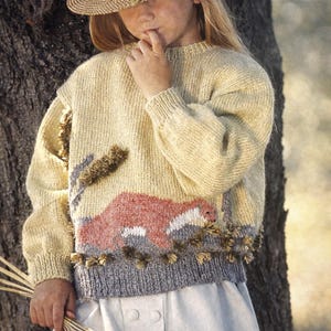 Könnte beinhalten: Ein Strohhut mit braunem Band und ein weizenfarbener Pullover mit Landschaftsdesign. Der Pullover hat einen grauen unteren Saum und dekorative Quasten. Die Person hält ein Bündel Weizen.