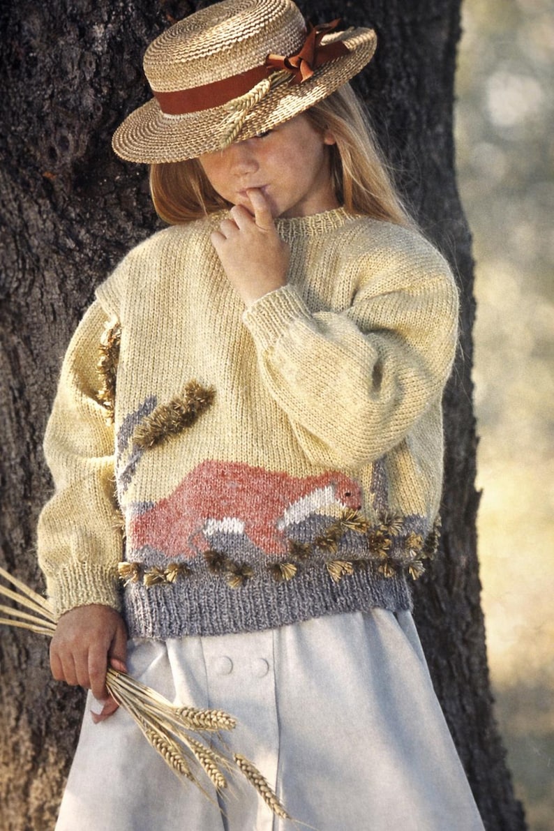 May include: A straw hat with a brown ribbon and a wheat-colored sweater with a landscape design. The sweater has a gray bottom hem and decorative tassels. The person is holding a bundle of wheat.