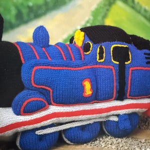 Pode incluir: Um comboio de brinquedo azul tricotado com detalhes vermelhos e amarelos. O comboio apresenta um topo preto, o número 1 amarelo e riscas vermelhas. As rodas são azuis com acabamento branco, numa base cinzenta e vermelha.