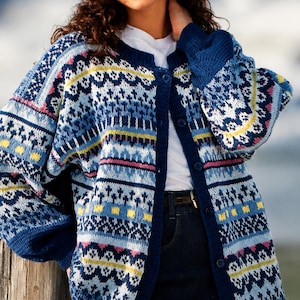 May include: A long-sleeve button-down cardigan featuring a complex knit pattern in shades of navy blue, light blue, white, yellow, and pink. The garment has a solid navy blue ribbed trim along the neckline, cuffs, and front button placket.