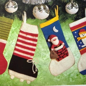 May include: Four knitted Christmas stockings hang from a garland. The stockings are red, green, white, and blue with various designs, including stripes, buttons, and a Santa Claus figure. Silver baubles are visible.