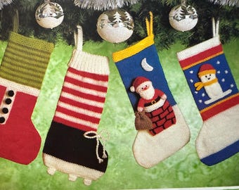 May include: Four knitted Christmas stockings hang from a garland. The stockings are red, green, white, and blue with various designs, including stripes, buttons, and a Santa Claus figure. Silver baubles are visible.