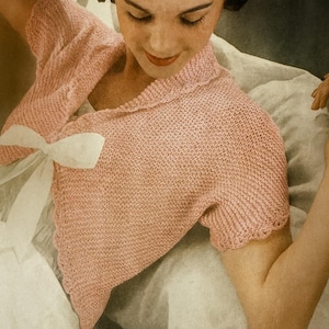 May include: A vintage-style, light pink knitted bolero with short sleeves and a white bow. The bolero has a scalloped edge and is made of a textured knit fabric. The garment is part of a vintage clothing collection.