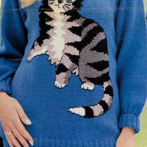 May include: A blue sweater featuring a large, detailed cat design. The cat is depicted in shades of black, gray, and white, with a striped pattern. The sweater has long sleeves and a ribbed hem and cuffs. The cat is centered on the front of the sweater.