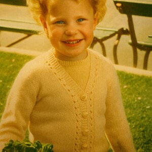 May include: A young child with curly blonde hair smiles, wearing a light yellow cardigan over a matching shirt. The cardigan has a V-neck and decorative button details. The child is holding green leafy plants.