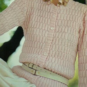 May include: A light pink, textured knit cardigan with a button-up front and long sleeves. The cardigan is cinched at the waist with a light-colored belt. A decorative necklace is worn at the neckline. The cardigan's knit pattern is a repeating rectangular design.