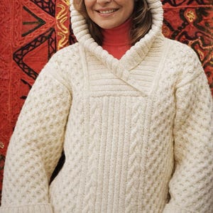 May include: A cream-coloured hooded sweater with a textured knit pattern. The sweater has a wide collar, long sleeves, and a relaxed fit. The background is a red and gold patterned textile.