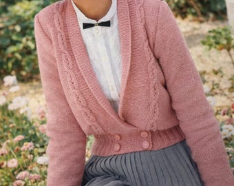 May include: A pale pink knitted cardigan with cable detailing on the sleeves. The cardigan features a shawl collar and two buttons. The model is wearing a white blouse with a black bow tie and a grey pleated skirt.