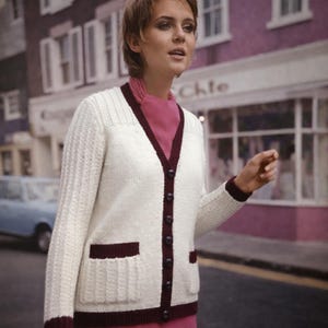 Op de afbeelding: Crèmekleurig vest met een V-hals en donker bordeauxrode bies. De trui heeft twee voorzakken en is aan de voorkant dichtgeknoopt. De mouwen en voorpanelen hebben een geribbelde textuur. Het vest wordt over een roze top gedragen.