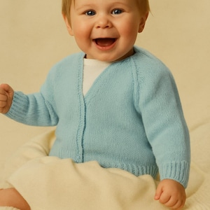 以下が含まれることがあります： 白いシャツの上に、Vネックとボタン留めの水色のカーディガンを着た笑顔の赤ちゃん。赤ちゃんはクリーム色の毛布の上に座っています。カーディガンはニットです。