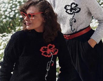 May include: Two women in floral sweaters. One wears a black sweater with a red flower, the other a grey sweater with a black flower. The woman on the left has red glasses and a red skirt. The woman on the right wears a red belt and black trousers.