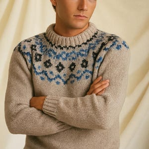 Könnte beinhalten: Ein beigefarbener Strickpullover mit Rundhalsausschnitt und langen Ärmeln. Der Pullover hat ein Muster in Blau und Schwarz um den Halsausschnitt und die obere Brust. Die Person auf dem Bild hat die Arme verschränkt.