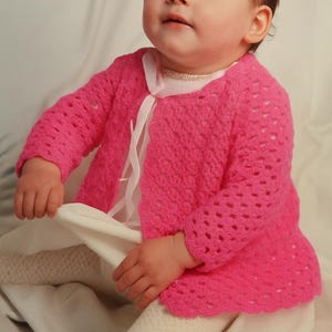 May include: A bright pink, open-weave baby cardigan with long sleeves. The cardigan is paired with white tights and a white dress with a ribbon tie. The baby is sitting on a white surface.
