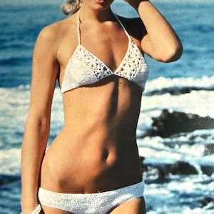 May include: A white crochet bikini. The halter-neck top has triangle cups, and the bottoms are low-rise. The bikini is made of textured, open-weave crochet fabric. The background shows a beach scene with blue water and rocks.