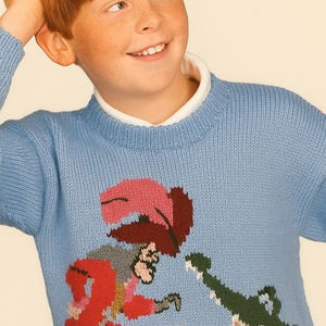 May include: A pale blue knitted jumper with a colourful pirate and crocodile design. The jumper has a crew neck and long sleeves. The pirate wears a red coat and a pink hat. The crocodile is green and brown.