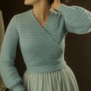 Op de afbeelding: Een lichtblauwe wikkel trui met een geschulpte halslijn en lange mouwen. De trui wordt gecombineerd met een lichtblauwe rok. De outfit heeft een vintage stijl, geschikt voor een retro of klassieke uitstraling.