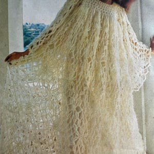 May include: A cream-coloured, open-weave crochet dress with long sleeves. The dress has a loose, flowing silhouette and a textured, fringed appearance. The crochet pattern features a combination of solid and open areas, creating a delicate, airy look.