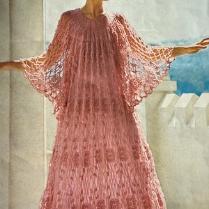 May include: A vintage, floor-length, pink crocheted dress with a cape-like top. The dress features a delicate open-weave pattern and floral detailing on the skirt. The model is standing outside with arms outstretched, showcasing the dress's flowing design.