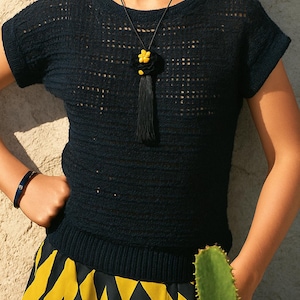 Puede incluir: Top de punto azul marino oscuro con cuello redondo y mangas cortas. El top se combina con una falda con estampado geométrico negro y amarillo. Se lleva un collar negro con colgante de flor y borla.