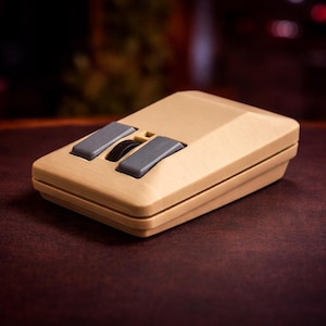 May include: A beige computer mouse with a rectangular body and two dark gray rectangular buttons positioned at the front. A small black scroll wheel is located between the two buttons. The device has a smooth, matte finish and a simple design.