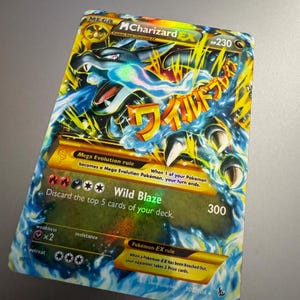 May include: A Mega MCharizard EX Pokémon trading card. The card features a blue and yellow dragon illustration with the text "Wild Blaze" and a 300 attack power. The card also includes the text "Mega Evolution rule".