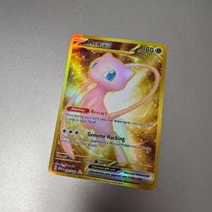 May include: A gold-colored Mew ex Pokémon trading card. The card features a pink Mew character with blue eyes and a long tail. The card has text describing the character's abilities and attacks.