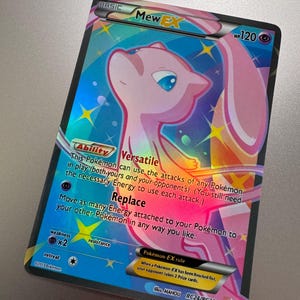 May include: A holographic Mew EX Pokémon trading card. The card features a pink Mew character with blue eyes against a colorful background. The card has text describing its abilities and rules, including "Versatile" and "Replace".