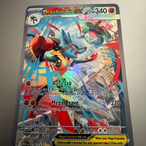 May include: A holographic Mega Lucario EX Pokémon trading card. The card features a blue and red Lucario character with the text "Aura Jab" and "Mega Brave". The card also includes the text "Mega Evolution EX rule" and "When your Mega Evolution Pokémon EX is Knocked Out, your opponent takes 3 Prize cards."