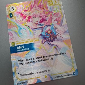 May include: A rectangular trading card featuring a pink-haired character named Ahri Inquisitive. The card displays a cost of 3, a power of 3, and text stating, When I attack or defend, give an enemy unit here -2 this turn, to a minimum of 1.