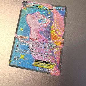May include: A holographic Pokémon trading card featuring Mew. The card has a pink and blue color scheme with sparkling accents. Text includes "Mew", "Versatile", and "Replace". The card is a basic Pokémon-EX rule card.