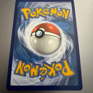 Puede incluir: El reverso de una carta coleccionable de Pok&eacute;mon. Muestra el logotipo de Pok&eacute;mon en amarillo y azul, y un dise&ntilde;o de Pok&eacute; Ball en el centro. La tarjeta tiene un borde azul y un fondo arremolinado.